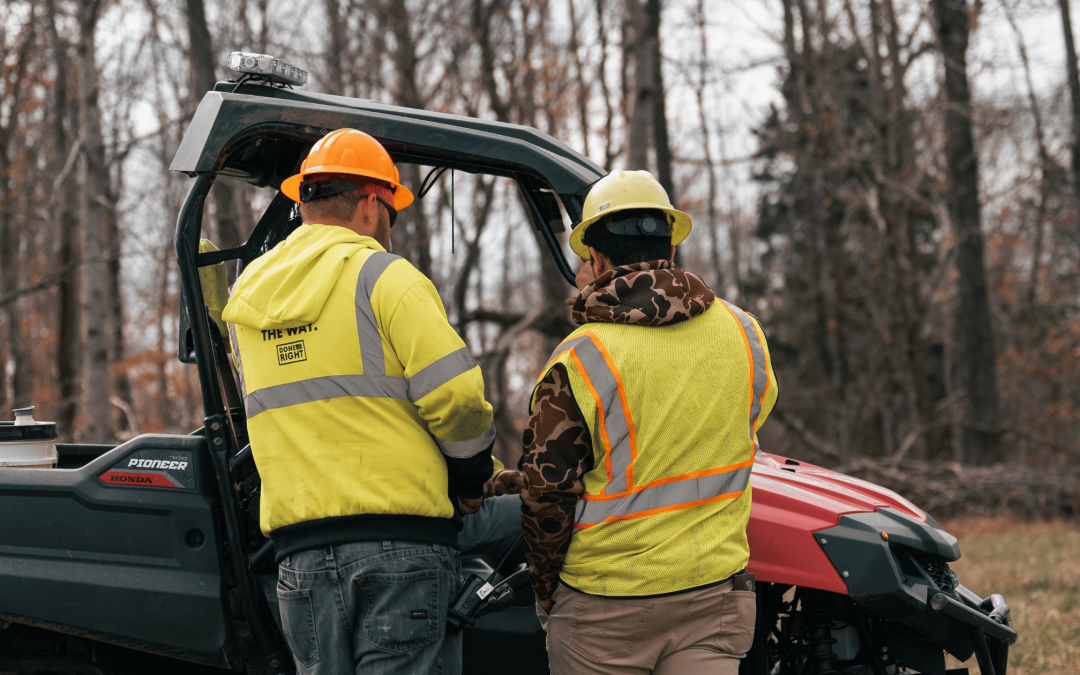 Two project managers wearing hard hats and safety vests for utility and industrial project management standing in front of side by side on a winter day with trees in the background