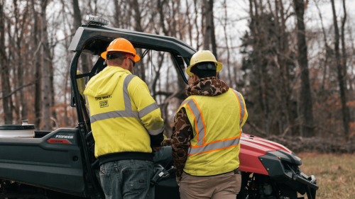 Two project managers wearing hard hats and safety vests for utility and industrial project management standing in front of side by side on a winter day with trees in the background